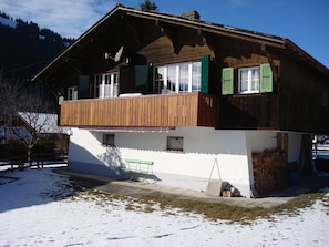 A two-story wooden house with a balcony, a small attached shed, and a bench outside.