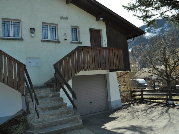 A two-story building with a wooden staircase, a garage, and a view of a snowy mountain.