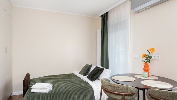 Et hotelværelse med en seng, et spisebord med stole, en vase med blomster og en aircondition.