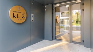 En moderne indgang med glasklar dør, digital elevatorpanel og et rundt skilt med bygningens nummer og adresse.