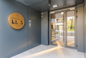 Ein moderner Eingangsbereich mit Glastür, digitaler Liftbedienung und einem runden Schild mit Hausnummer und Adresse.
