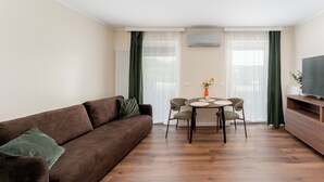 En stue med en brun sofa, et spisebord med stole, et fjernsyn på et træmøbel og en radiator.