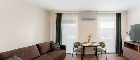 Un salon avec un canapé marron, une table à manger avec des chaises, un téléviseur posé sur un meuble en bois et un radiateur.
