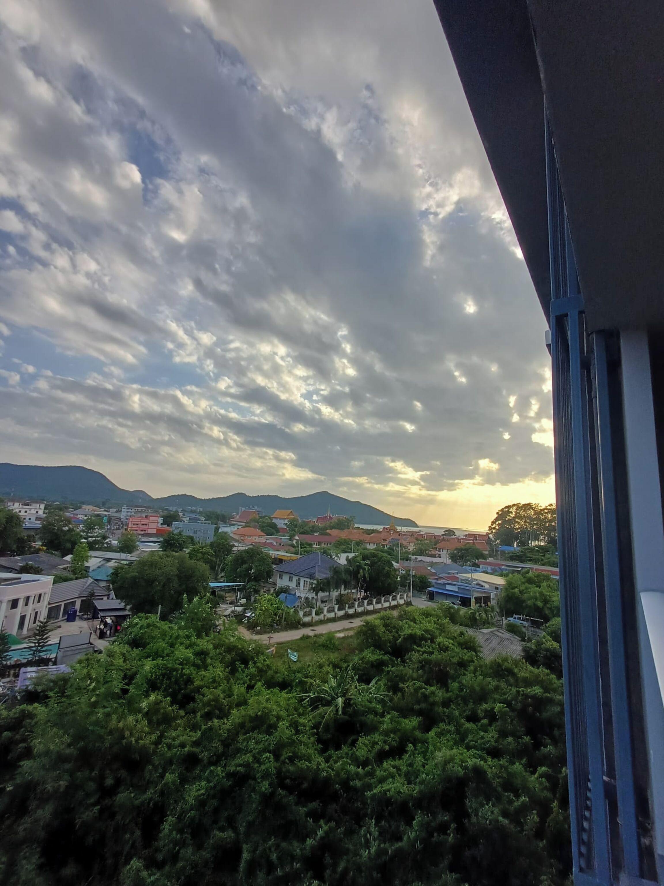 A view of a town with buildings and greenery, a clear sky with clouds, and mountains in the distance.