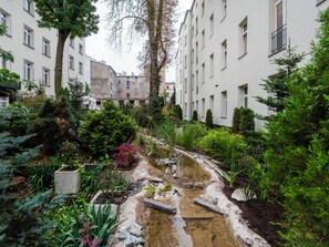 Um pátio com um pequeno elemento aquático, rodeado por vegetação exuberante e edifícios modernos.