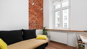 Ein modernes Wohnzimmer mit einer schwarzen Couch, gelben Kissen, einer Ziegelwand, einem Fenster mit Jalousien und einem Essbereich mit gemusterten Stühlen.