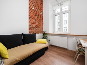 A modern living room with a black sofa, yellow cushions, a brick wall, a window with blinds, and a dining area with patterned chairs. - Kamienica Rewolucji 23 Mezzanine Studio (Łódź)