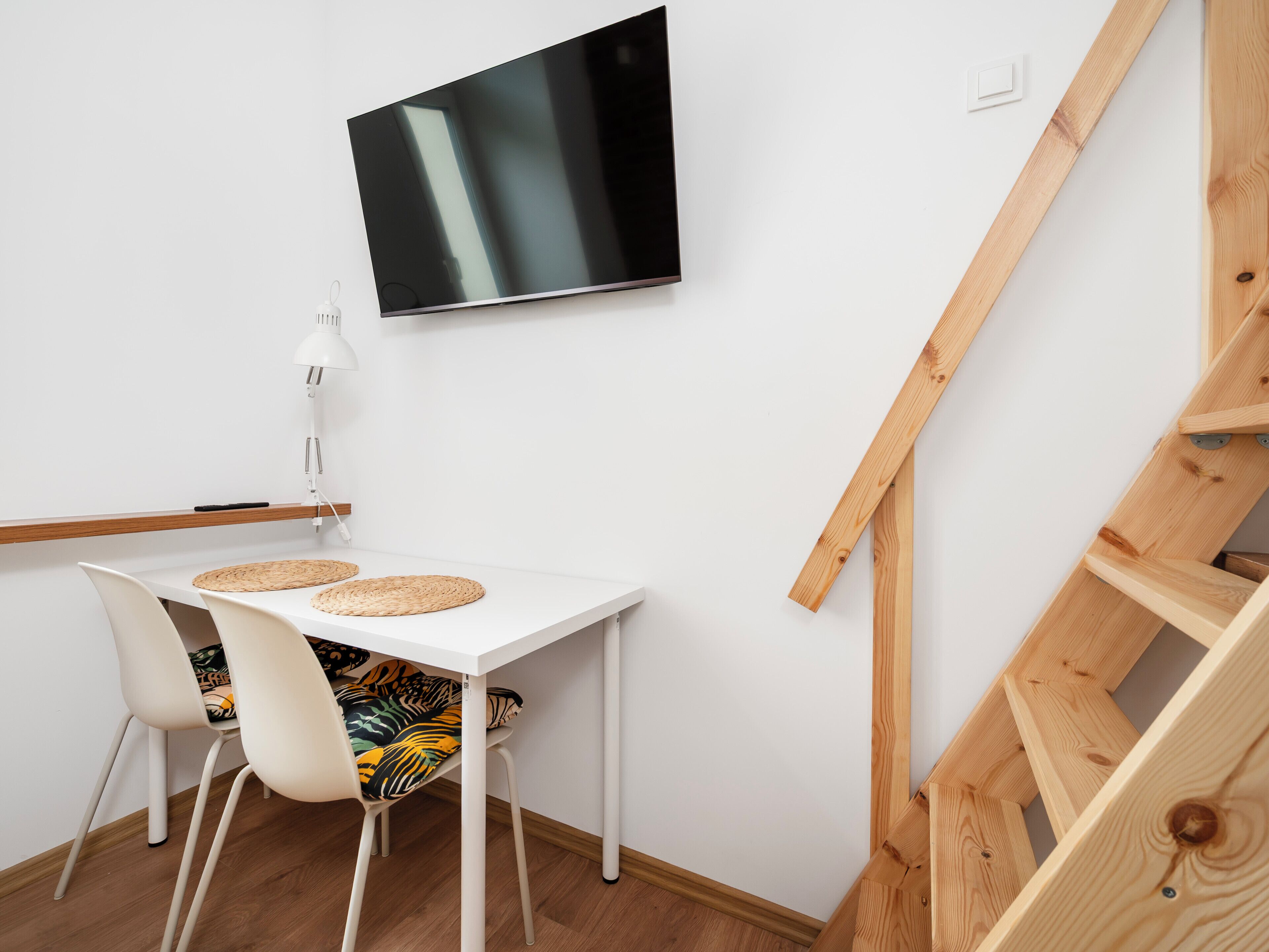 Une pièce moderne avec une table blanche, deux chaises, un téléviseur à écran plat, une lampe et un escalier en bois.