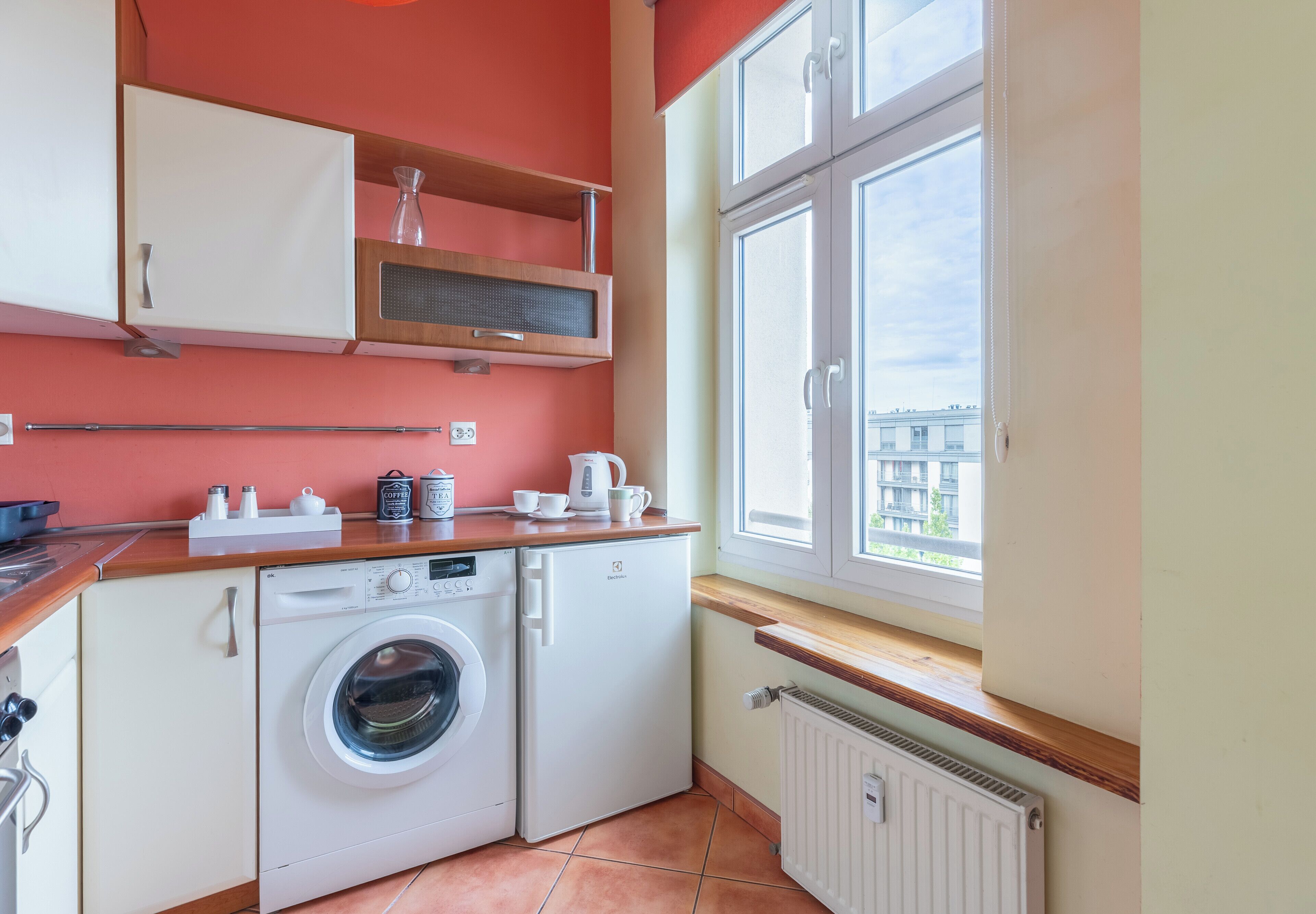 Une cuisine compacte équipée d’un lave-linge, d’une machine à café et d’une fenêtre donnant sur des immeubles.