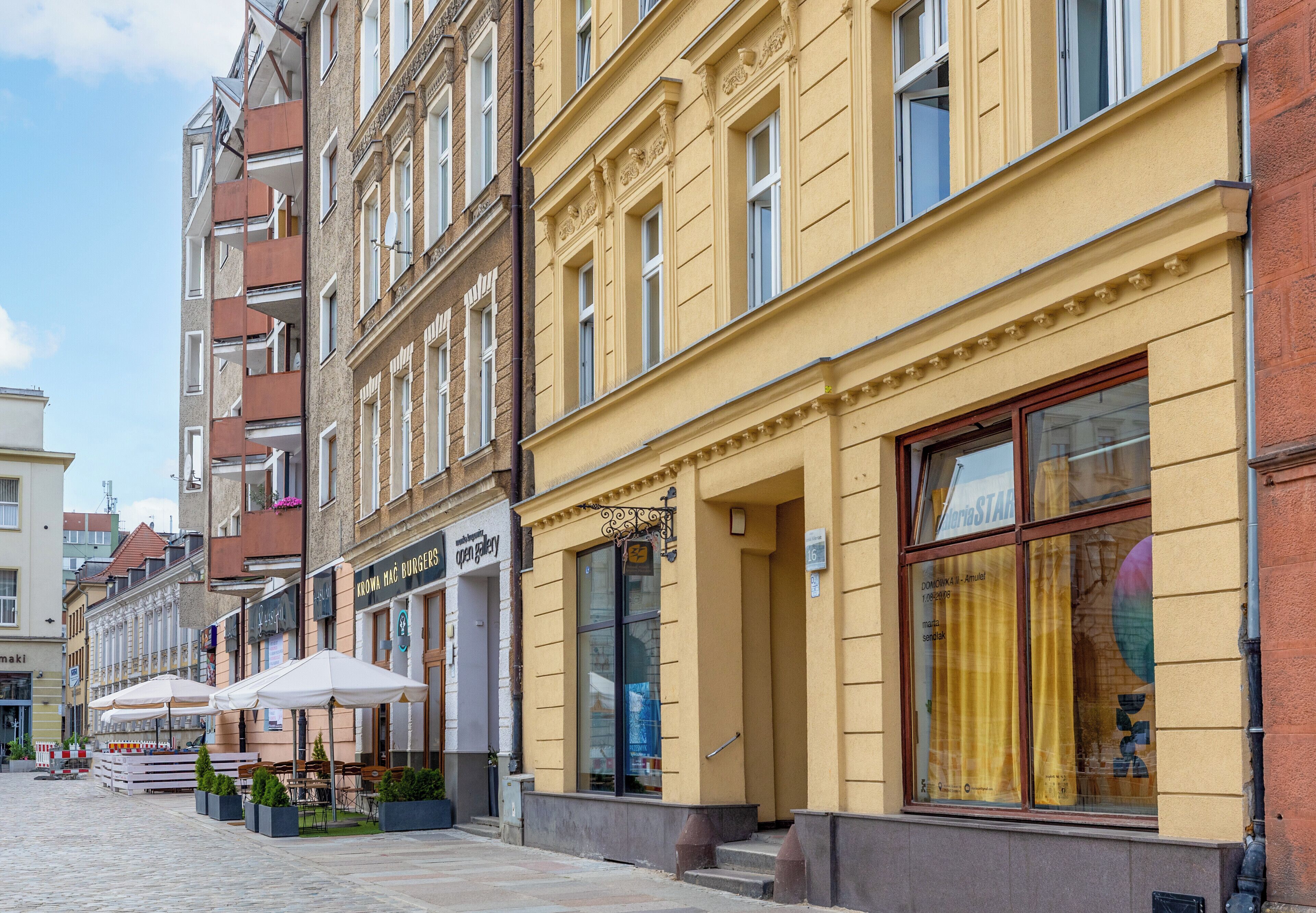 Eine Straße mit Gebäuden, Außenbestuhlung und blauem Himmel.