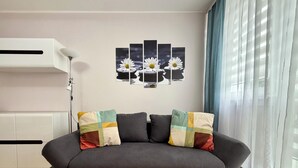 Ein modernes Wohnzimmer mit einem grauen Sofa, bunten Kissen, einem Wandbild mit Margeriten und einem weißen Heizkörper.