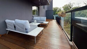 Uma área de estar moderna ao ar livre, com um deck de madeira, sofás cinzas e uma cesta suspensa.