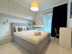 A modern bedroom with a bed, bedside lamp, and a window with blinds. - Kępa Mieszczańska Spacious Apartment (Wrocław)
