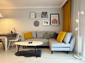 A modern living room with a grey sofa, a round coffee table, and a fluffy rug. There are framed pictures on the wall, a clock, and a desk with a lamp. - Kępa Mieszczańska Spacious Apartment (Wrocław)