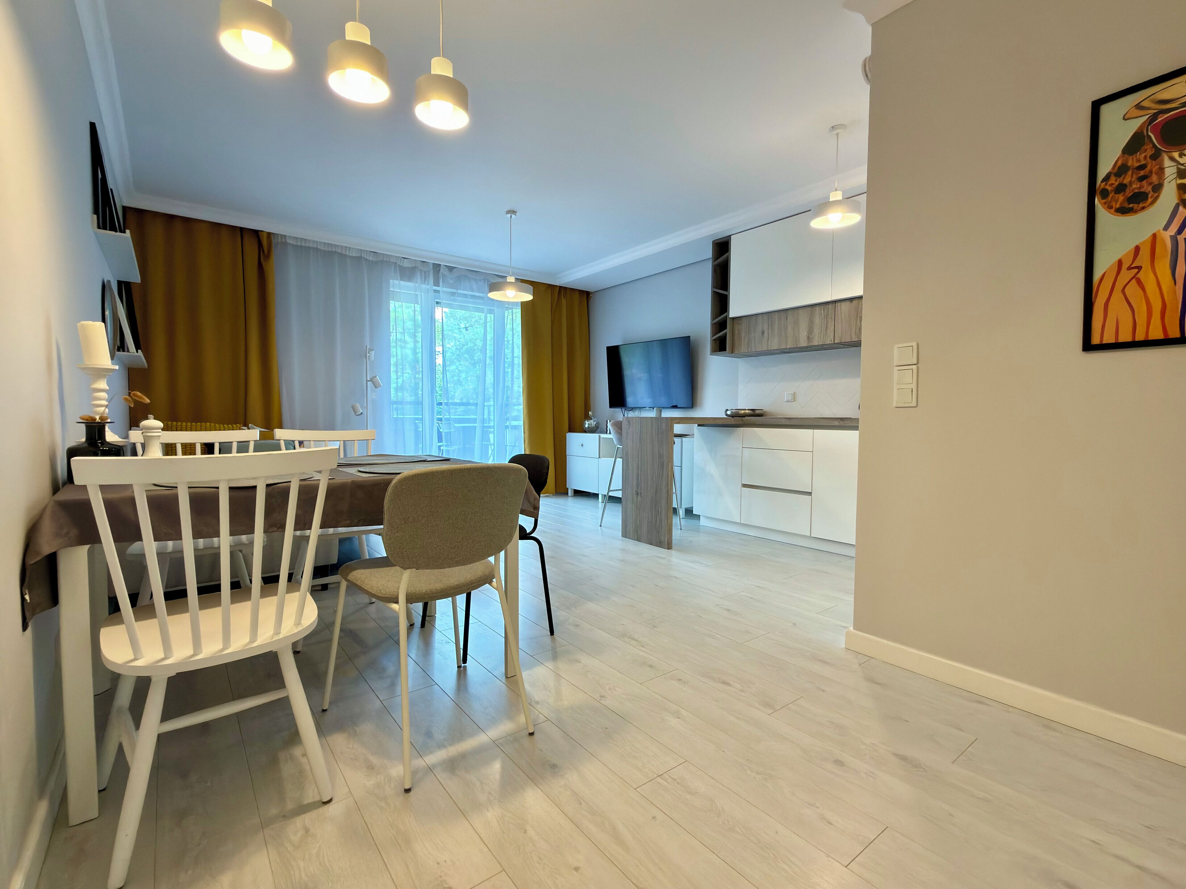 A modern kitchen with a flat-screen TV, a dining area with a white table and chairs, and a large window with curtains.