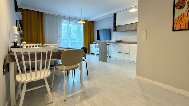 Uma cozinha moderna com televisão de tela plana, uma área de refeições com mesa e cadeiras brancas, e uma janela grande com cortinas.