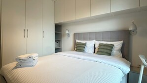 Cama bem arrumada com edredom branco e almofadas, mesa de cabeceira branca e guarda-roupa com prateleira.