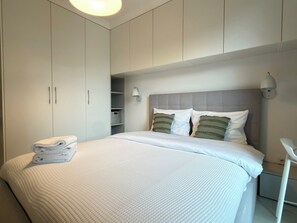 Ein ordentlich bezogenes Bett mit weißer Bettdecke und Kissen, ein weißer Nachttisch und ein Kleiderschrank mit Ablagefläche.