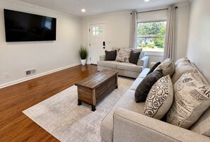 A living room with a sofa, a coffee table, a TV mounted on the wall, and a window with curtains. - Spacious 3BR Home Near Truist Park and I-75 (Marietta)