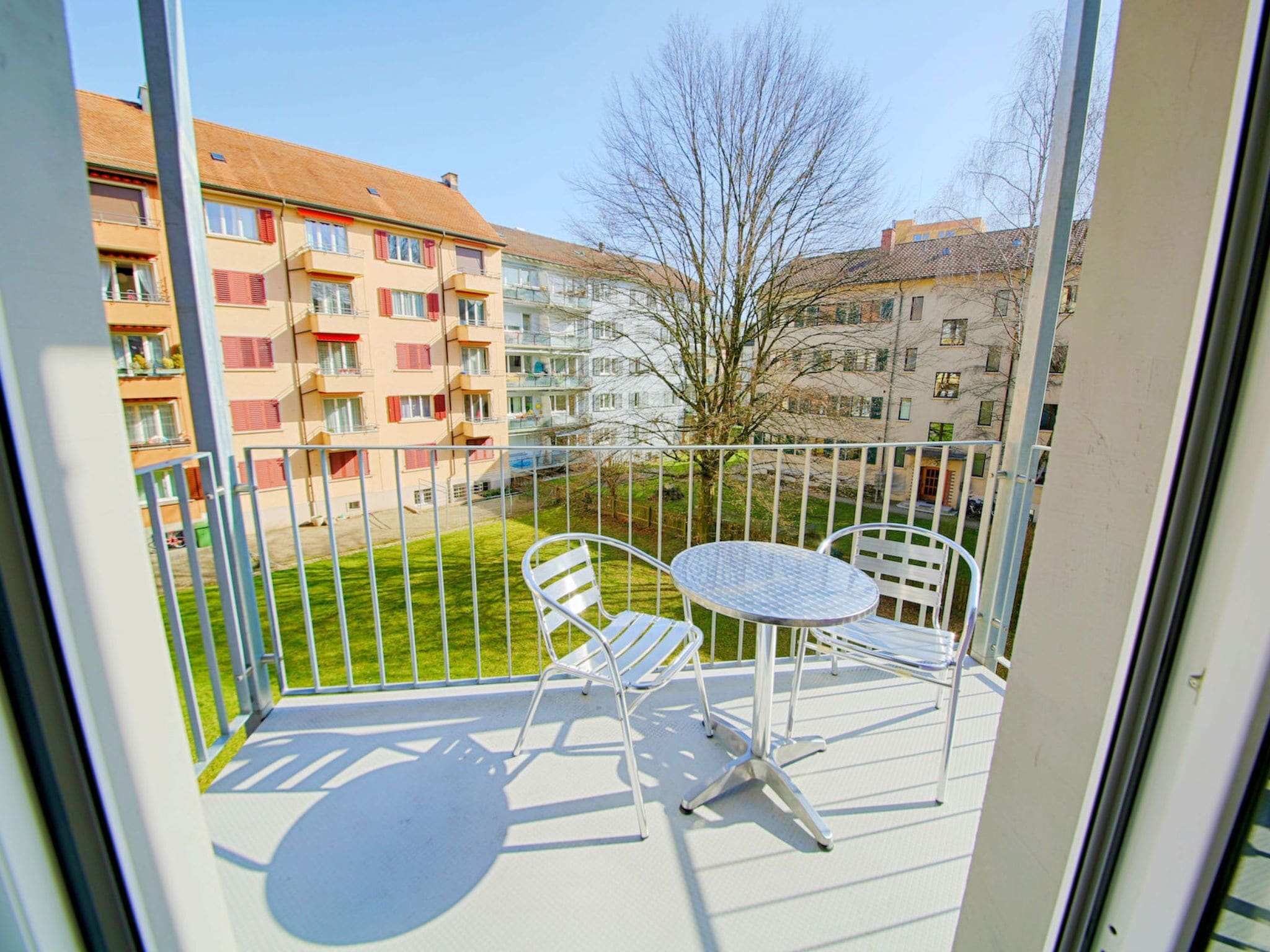 A balcony with a metal table and two chairs, overlooking a grassy area and buildings.