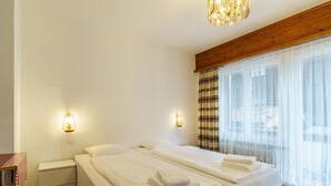 Quarto de hotel com cama de casal, mesas de cabeceira, luminária e uma janela grande com cortinas.