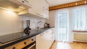 Ett modernt kök med spis, ugn och diskho, trägolv och ett fönster med tunna, genomskinliga gardiner.