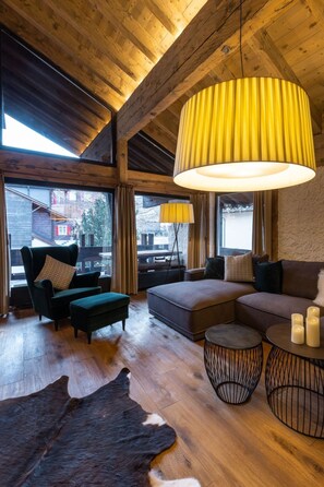 Ein geräumiges Wohnzimmer mit hoher Decke, Holzbalken, einem großen Fenster und einer modernen, gelben Pendelleuchte.