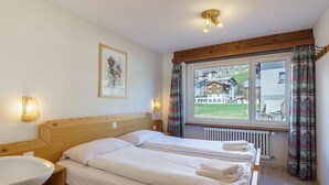 A hotel room with two beds, a wooden headboard, a bedside table with a white sink, and a window with curtains. - Haus Alfa by Interhome (Saas-Fee)