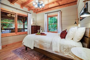 Une chambre à coucher comprenant un lit, une table de chevet, une commode, une fenêtre donnant sur un paysage verdoyant et un lustre.