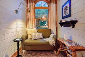 A cozy room with a green sofa, a small table with a lamp, and a window with a view of greenery. - The Smoky Edit | 3 Decks | Hot Tub | Pool Table | 2 Fire Pits (Gatlinburg)