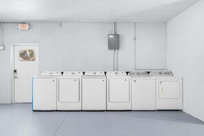 A laundry room with a row of white washers and dryers, a door with a window, and an exit sign on the wall. - Poolside Fun, Pickleball & Coastal Adventures Await in Fort Myers - Jefferson Palms 121 - Roelens (Fort Myers Beach)