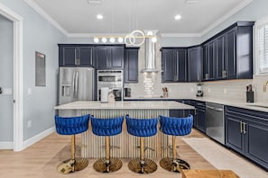 A modern kitchen with dark cabinetry, a stainless steel refrigerator, an oven, a coffee maker, and a bar stool area. - Poolside Fun, Pickleball & Coastal Adventures Await in Fort Myers - Jefferson Palms 121 - Roelens (Fort Myers Beach)