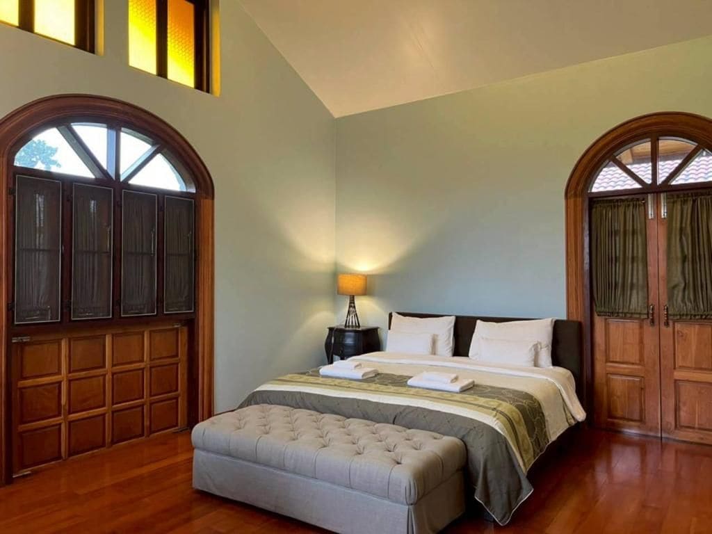 Un dormitorio con una cama grande, una mesita de noche y un banco. Hay dos ventanas amplias con marcos de madera y cortinas.