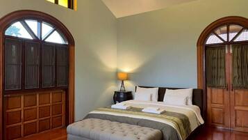 Un dormitorio con una cama grande, una mesita de noche y un banco. Hay dos ventanas amplias con marcos de madera y cortinas.