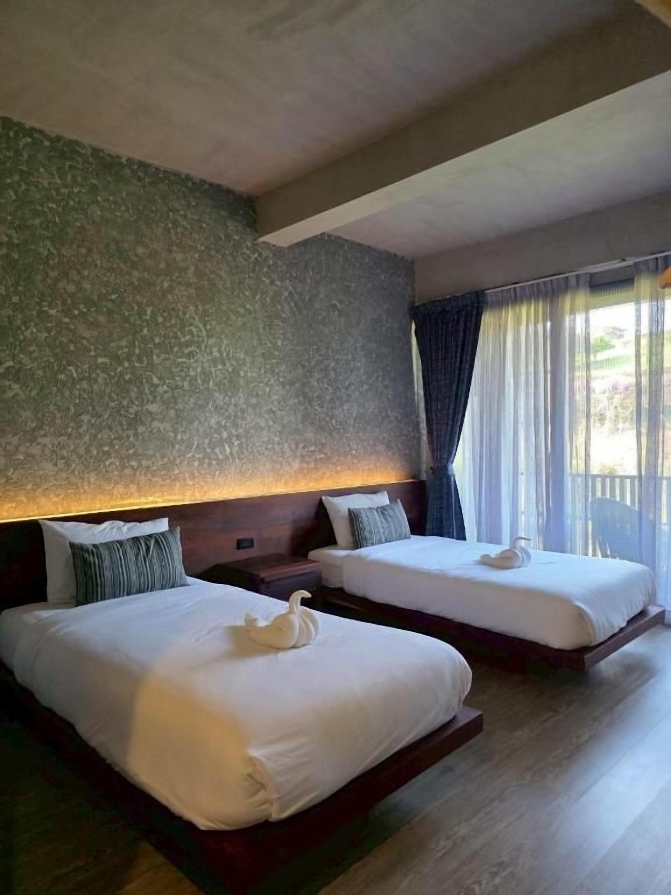 Quarto de hotel com duas camas, parede texturizada, piso de madeira e janela com cortinas.