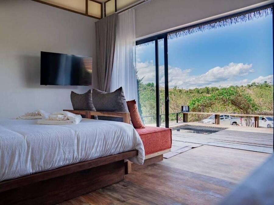 A bedroom with a large bed, a flat-screen TV, a view of the outdoors through a sliding glass door, and a balcony with a wooden deck.