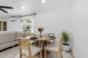 Een eetgedeelte met een ronde tafel, vier stoelen, een plant in pot en een raam met jaloezieën.
