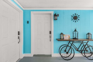 A hallway with a white door, a turquoise wall, a bicycle, and a small table with a sign. - Poolside Fun, Pickleball & Coastal Adventures Await in Fort Myers - Jefferson Palms 125-7b - Roelens (Fort Myers Beach)