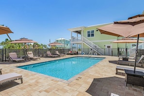 A swimming pool with lounge chairs and umbrellas, surrounded by a fence and a building with a balcony. - Poolside Fun, Pickleball & Coastal Adventures Await in Fort Myers - Jefferson Palms 125-7b - Roelens (Fort Myers Beach)