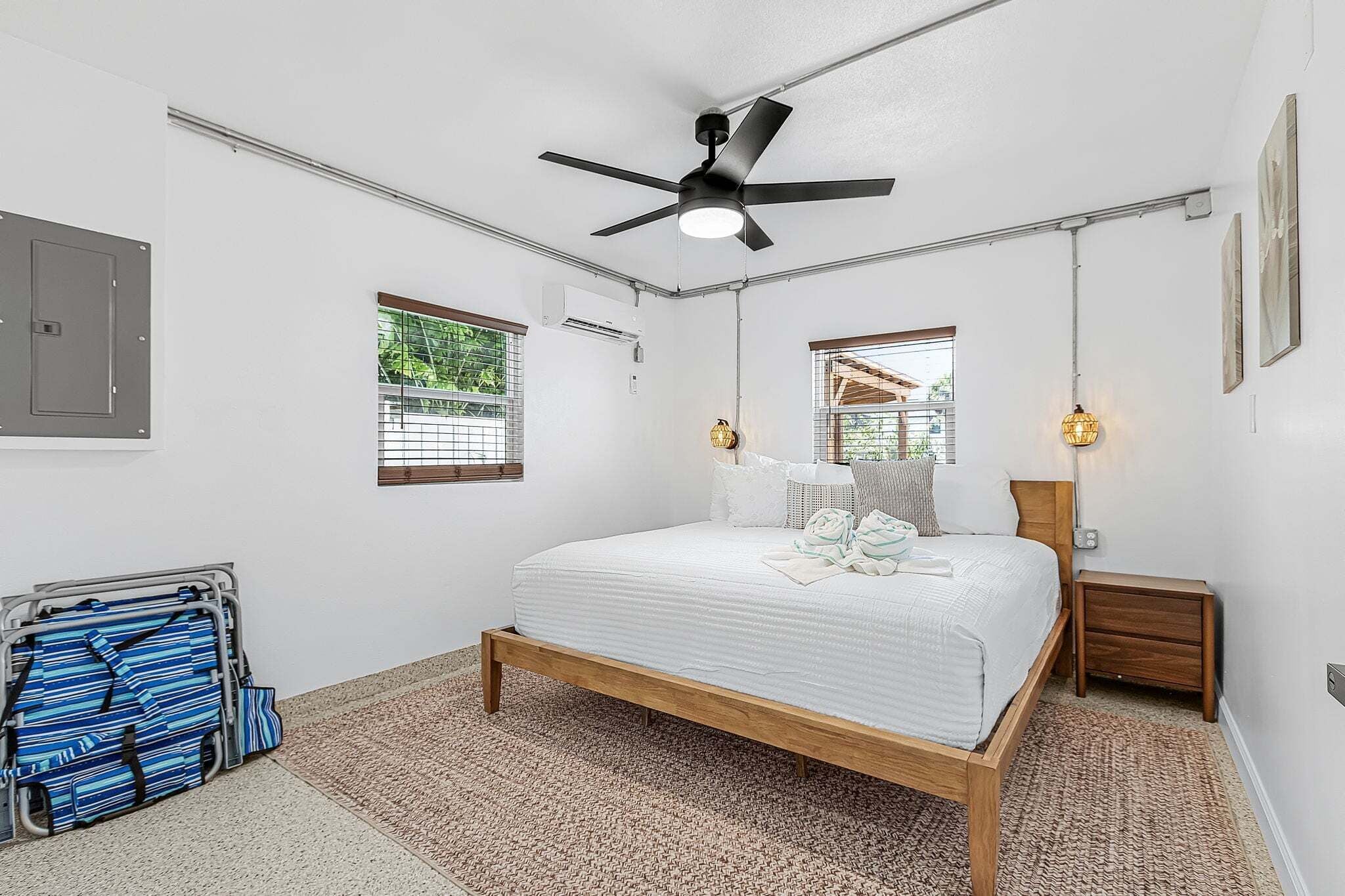Ein Schlafzimmer mit einem Bett, Deckenventilator, zwei Fenstern mit Jalousien, einer Nachttischlampe, einem Nachttisch und einem Koffer.