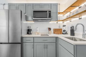 A modern kitchen with grey cabinetry, a stainless steel refrigerator, a built-in microwave, a coffee maker, a blender, and a wooden cutting board. - Poolside Fun, Pickleball & Coastal Adventures Await in Fort Myers - Jefferson Palms 125-8 - Roelens (Fort Myers Beach)