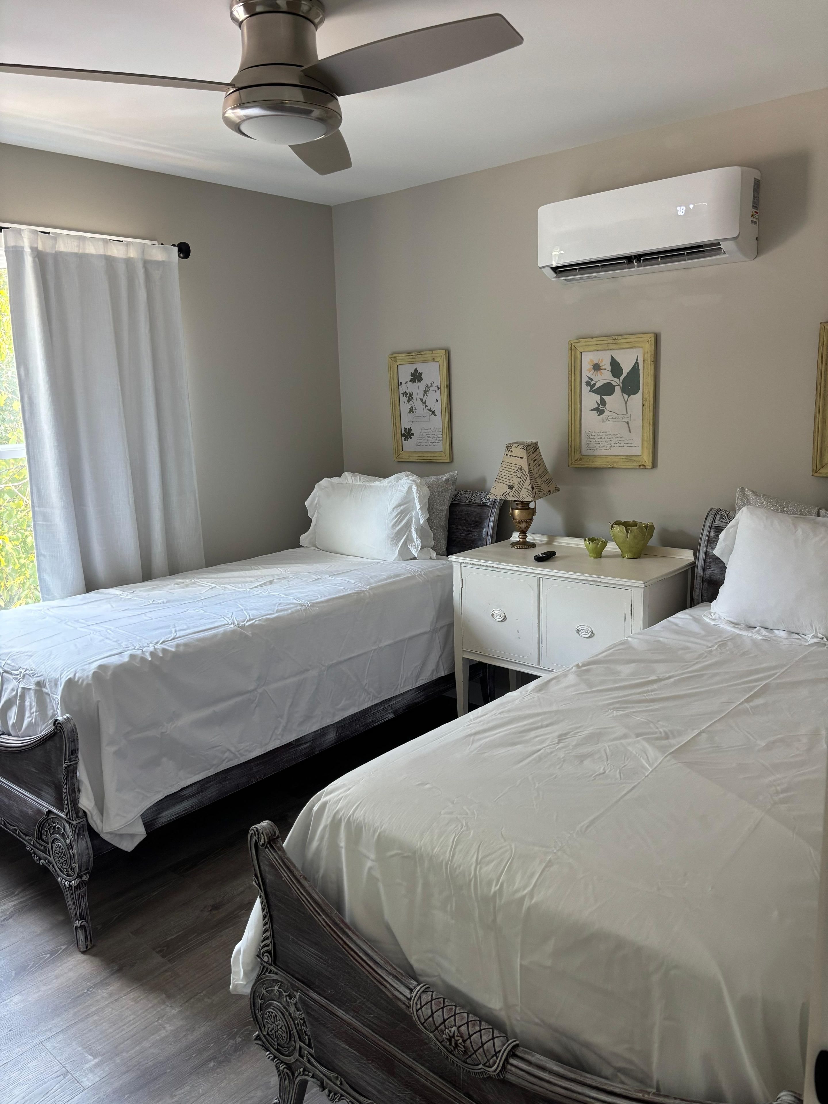 Habitación de hotel con dos camas, ventilador de techo, unidad de aire acondicionado, mesita de noche con lámpara e impresiones botánicas enmarcadas en la pared.