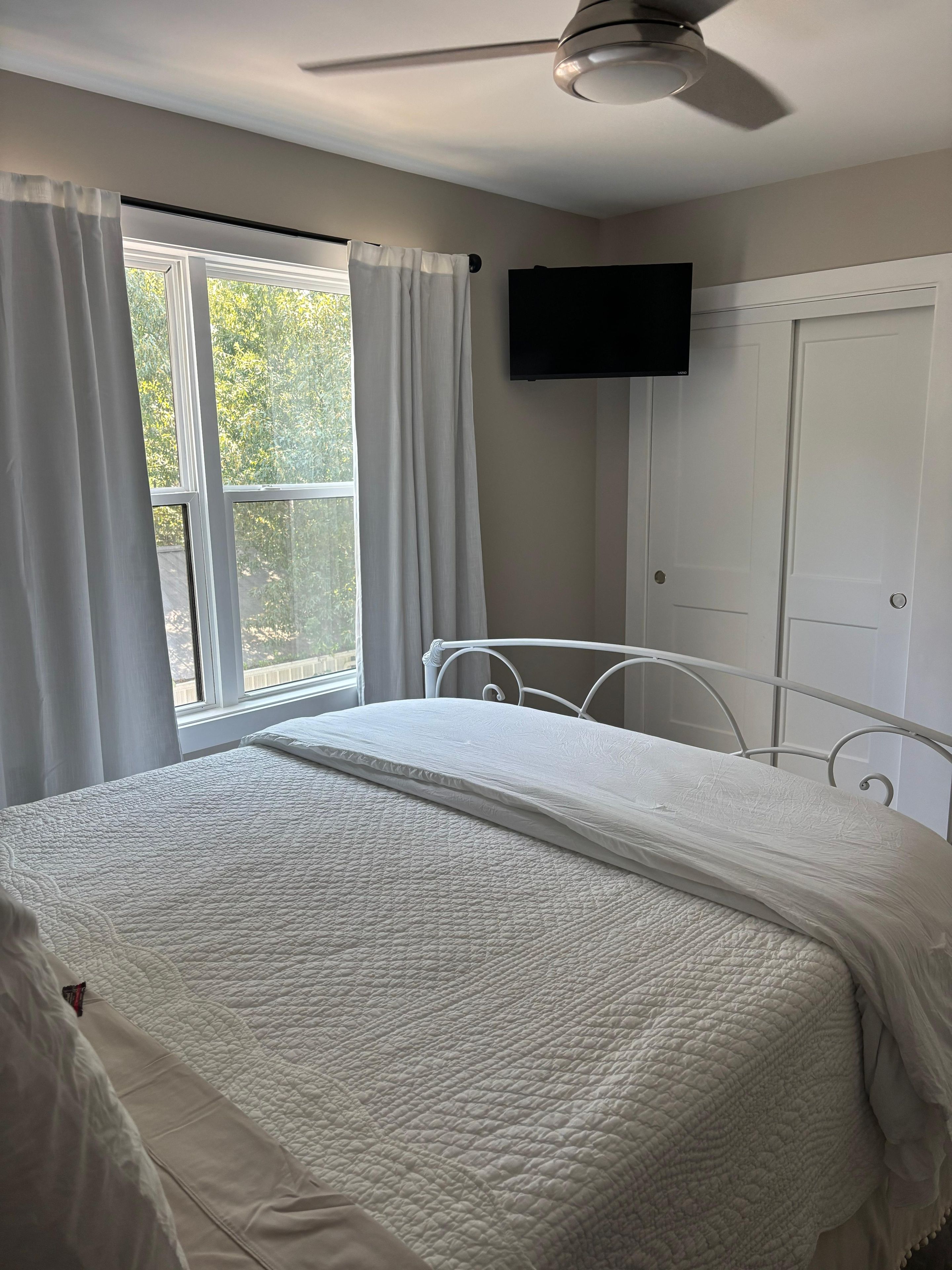 A bedroom with a bed, a ceiling fan, a window with curtains, and a television on the wall.