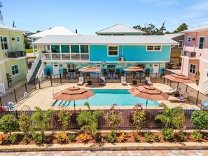 A pool area with lounge chairs and umbrellas, a multi-story building with a blue facade, and a landscaped garden with various plants. - Poolside Fun, Pickleball & Coastal Adventures Await in Fort Myers - Jefferson Palms 127 - Roelens (Fort Myers Beach)
