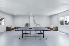 A room with two ping pong tables, wicker furniture, a potted plant, and large windows. - Poolside Fun, Pickleball & Coastal Adventures Await in Fort Myers - Jefferson Palms 127 - Roelens (Fort Myers Beach)