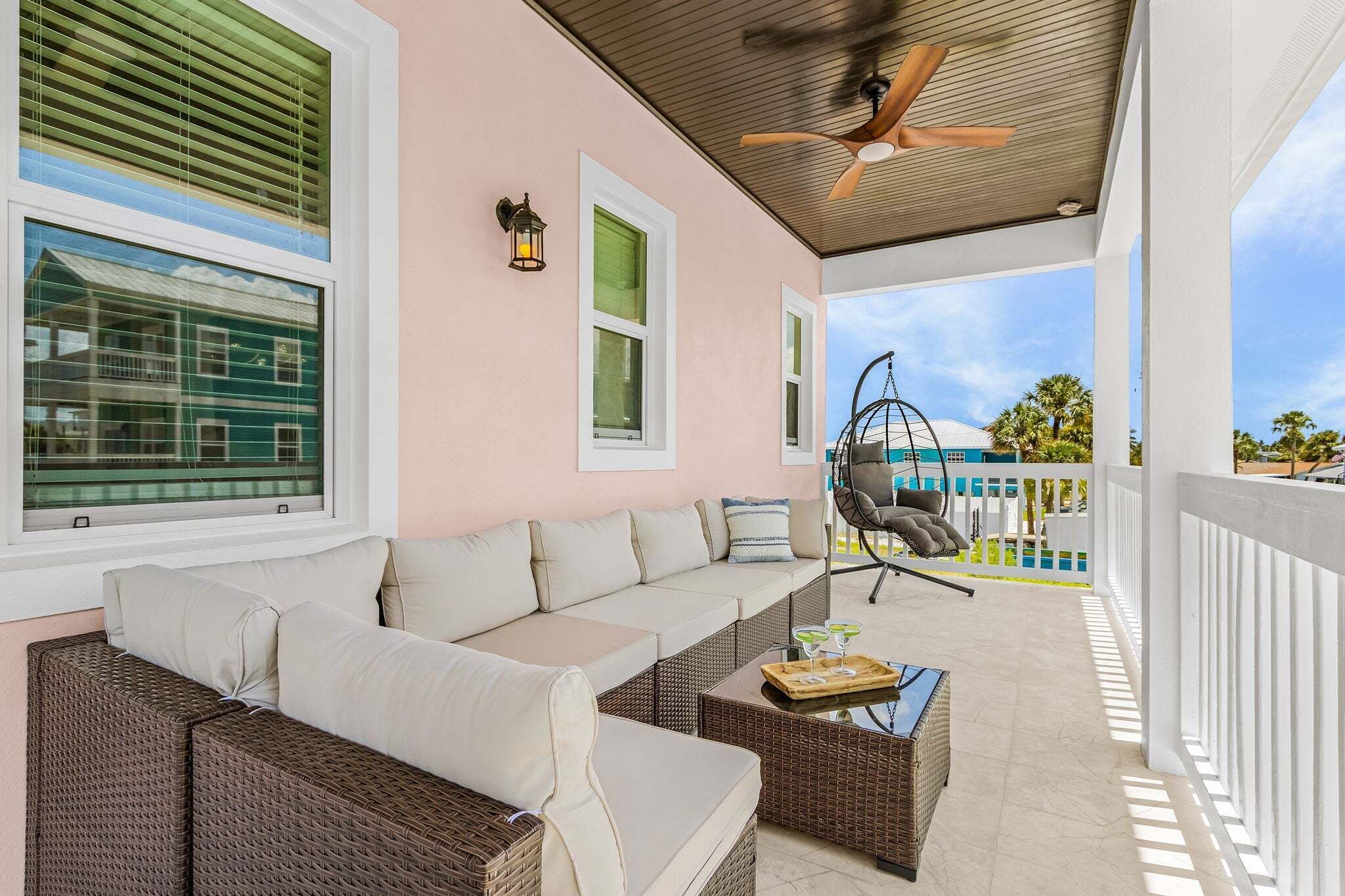A covered outdoor seating area with a sofa, wicker furniture, a hanging chair, and a ceiling fan.