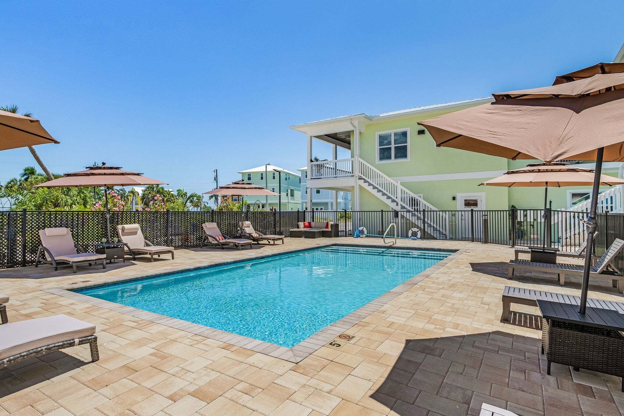 A swimming pool with lounge chairs and umbrellas, surrounded by a fence and a building with a balcony.