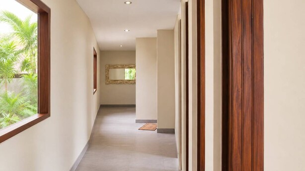A corridor with a wooden door, a mirror, and a window with a view of greenery.