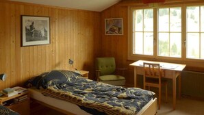 Um quarto com uma cama, uma escrivaninha, uma cadeira e duas janelas.