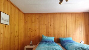 A room with wooden panel walls, a bed with blue bedding, a nightstand with a lamp, and a ceiling fan. - Schwendli Appartement/Fewo, Dusche, WC 3-Zimmerwohnung by Interhome (Kandersteg)